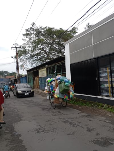 A street scene in Kecamatan Kutawaringin, Indonesia (latitude: -6.974, longitude: 107.547). A person in red clothing is walking on the street, facing towards the left side of the image. A person is riding a motorcycle on the street, facing towards the left. A grey car is stopped in the middle of the street, facing towards the left. A motorcycle is parked on the left side of the street. A cart loaded with various objects including a plastic bucket, a broom, and other household items, is being pulled by a man on the right side of the street. The man is obscured from view. There are power lines running across the top of the image.  A building with a black metal door is on the right side of the image. A tree is on the left side of the image, with the top of the tree branches out of frame. The ground is paved asphalt, and the street has cracks and potholes.  The sky is overcast and grey.
