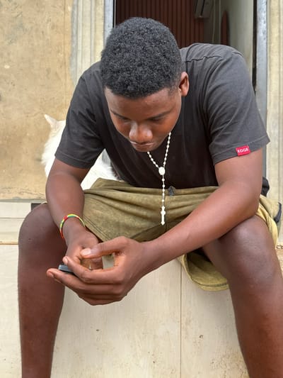A young person with dark skin and short, curly black hair is seated on a light-colored tiled floor. They are wearing a dark grey t-shirt and olive green shorts. A white beaded necklace hangs around their neck, and a colorful beaded bracelet in red, yellow, and green is visible on their right wrist. Their right hand holds a small dark object, possibly a mobile phone, and their gaze is directed downwards, suggesting focus or contemplation.

The person's posture is relaxed, with one leg bent and the other extended. In the background, elements suggest an indoor or sheltered outdoor setting, featuring what appears to be a metallic door frame or window, a textured wall, and a white air conditioning unit. A small, white furry object, possibly a pet like a dog, is partially visible near the person's left side.

The lighting is bright and even, indicating it is daytime. The overall mood is calm and neutral. A notable detail is the visible red label on the left sleeve of their t-shirt, which reads "EDGE" in white text. The scene is located in Ikot Ekpene, Nigeria.