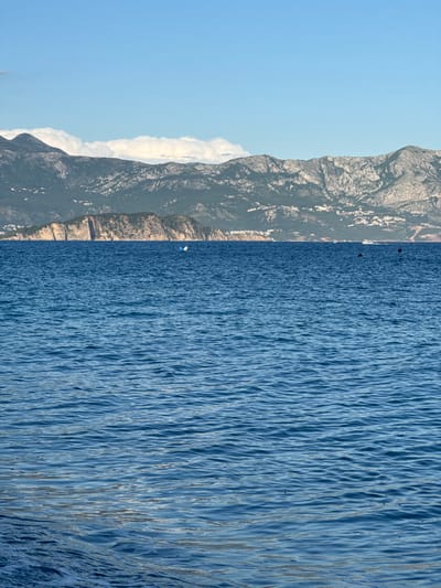 The image shows a calm blue sea with distant mountains. The sky is clear and blue with a few white clouds. In the middle ground, a rocky island is visible, with some buildings on its slopes. A white and blue object, possibly a buoy or a small boat, is in the water near the island. Further out, a white boat with people on it is moving across the water. The scene is a coastal landscape near Prijevor, Montenegro.