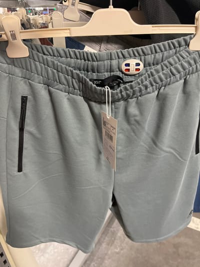 This media file is a close-up shot of a pair of light blue-grey sweatpants hanging on a clothing rack. The sweatpants have an elastic waistband and zippered pockets. A small white tag with red and blue accents is attached to the waistband. Another white tag, with visible text including a barcode and sizing information, is hanging from a string on the sweatpants. The background shows other clothing items on hangers and a blurred store environment. The image appears to have been taken indoors, likely in a retail setting, given the clothing racks and the general ambiance. No people are visible in the frame. The lighting suggests it is daytime. The text on the visible tag is mostly illegible due to the angle and resolution, but "M" is clearly visible indicating the size. The overall impression is a product shot focusing on the details of the garment.