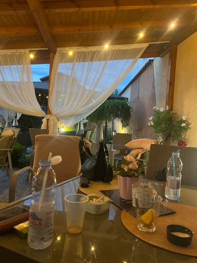 This image depicts a cozy outdoor seating area, likely a patio or garden at night, located in Dragoman, Bulgaria. The scene is illuminated by numerous warm yellow string lights that cast reflections on a clear, glass-topped table in the foreground.
On the table, various objects are arranged: a light brown woven straw hat, a black handbag with visible gold hardware, and a pink potted plant bearing light-colored flowers. Beverages include a clear glass mug containing a slice of lemon or fruit, and two plastic water bottles, one with a blue straw. The brand name "EDELA" is visible on one of the water bottles. Additionally, two clear plastic cups and a small black bowl with a light-colored substance are present on the table. A light brown, ribbed chair cushion is also visible.
The setting features wooden beams and structures, possibly part of a pergola or roof support, from which the string lights are strung. White, translucent curtains or drapes are tied back, framing the view into a garden or courtyard. In the background, dark green trees and shrubs are visible under a twilight or night sky, along with another large potted plant showcasing white and red blooms. The ambiance is inviting and relaxed, characteristic of an evening gathering or meal. No people are visible in the image.