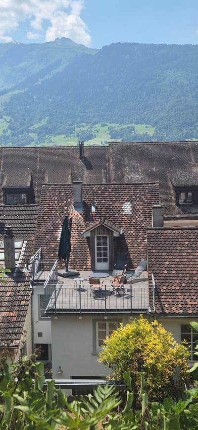 The image shows a rooftop scene in Sargans, Switzerland, with mountains in the background. The foreground features a terrace with outdoor furniture, including chairs, a table, and an umbrella. The terrace is surrounded by a metal railing. The roofs of adjacent buildings are visible, covered in reddish-brown tiles. A small wooden structure with a window and a door is present on the roof. In the distance, green hills and mountains are visible under a blue sky with some clouds. Lush green foliage is present at the bottom of the image, obscuring part of the lower buildings.