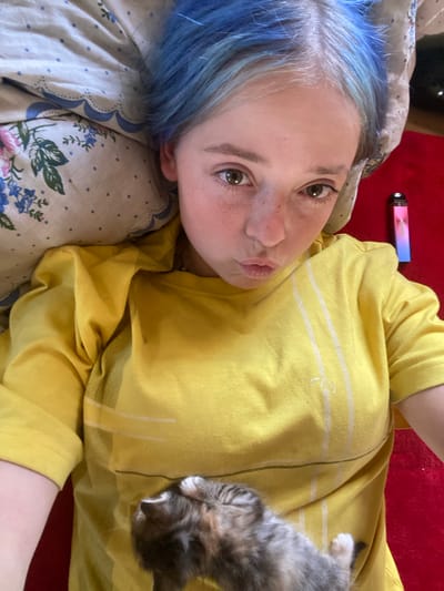 A close-up, low-angle shot shows a person with bright blue hair lying on their side, looking at the camera. They have fair skin with freckles and are puckering their lips. The person is wearing a bright yellow t-shirt with white stripes and a logo. A small, fluffy kitten with grey, brown, and white fur is nestled on the person's chest. The person is lying on a patterned blanket with blue and white floral designs, and to the right, a red surface is visible, possibly a carpet or bedspread. In the background, near the top left corner, a rectangular object with pink and blue light is visible, possibly an electronic device. The lighting suggests daytime indoors. The overall mood appears relaxed and affectionate due to the presence of the kitten. There is no visible text.