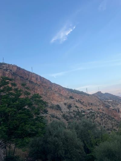 The image shows a mountainous landscape with rocky cliffs and sparse vegetation. There are power lines running along the mountainside. Some trees and bushes are visible in the foreground. The sky is blue with some wispy clouds. The location is likely near Bin El Ouidane, Morocco, based on the arid landscape and the presence of power infrastructure associated with the dam and reservoir in that area. No people are visible.