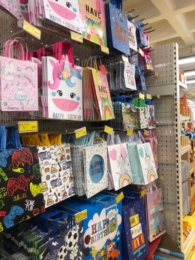 This media file presents a well-stocked retail display of numerous gift bags, likely intended for birthdays and other celebrations, within a store located in Marousi, Greece. No people are visible in the image, focusing entirely on the merchandise.
The gift bags are neatly arranged on multi-tiered, white metal shelving units. They feature a diverse range of colorful designs and themes. Many bags explicitly display "HAPPY BIRTHDAY" greetings. Other prominent designs include unicorns with phrases like "HAVE magical" and "dreams," cartoon elephants under rainbows, construction vehicles, footballs, and video game controllers with text such as "LETS PLAY," "SOME FUN," and "GAME." The bags come in various sizes and colors, with handles in coordinated or contrasting hues like pink, blue, green, and rainbow stripes.
Yellow price tags are visible on the shelving units, with some tags showing prices such as "79 A" and "1.00 A," indicating the cost of the items. The overall setting is an indoor commercial space, brightly illuminated by artificial lighting, typical of a discount store or a party supply shop. In the background, additional shelving units filled with other products can be seen, suggesting a large retail environment.