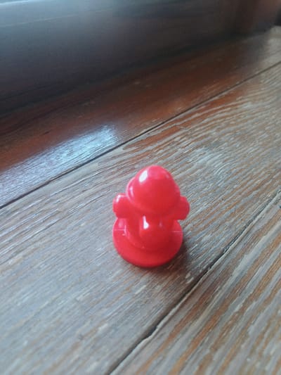A red, plastic duck figurine sits on a weathered wooden surface. The duck is small and appears to be a bath toy. The wooden surface has visible grain and appears to be made of planks. The lighting suggests an indoor environment, with a shadow cast from an unseen object. No specific location in the city of Odesa, Ukraine can be determined from the image.