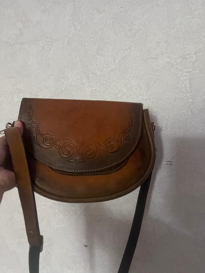 A person's hand is holding a brown leather crossbody bag. The bag has a flap closure with ornate embossed designs and a dark strap. The bag is positioned against a textured white wall, possibly in an indoor setting. The overall lighting suggests a dimly lit environment. There is no indication of the specific location being Şamaldı-Say, Kyrgyzstan.
