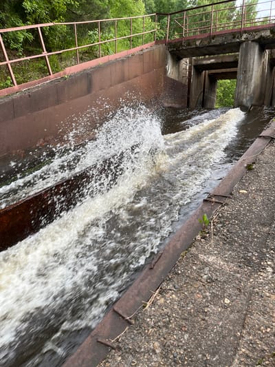 The image shows a spillway with water flowing through it. The spillway is constructed of concrete and rusted metal. There is a metal railing on the left side of the spillway, and trees and foliage are visible in the background. The water is turbulent and appears to be flowing rapidly. The scene is outdoors, and the weather appears to be overcast. The location is identified as Chernushka, Russia.