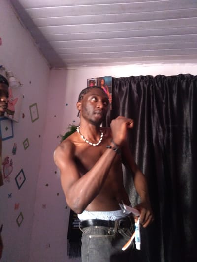 The image depicts a young man with braided hair, shirtless, wearing a necklace and jeans, holding a tube of toothpaste and a toothbrush. He is standing in a room with a patterned wall, possibly decorated with cartoon characters and geometric shapes. A black curtain hangs in the background, and a fluorescent light fixture is visible on the ceiling. The overall impression is that of a candid snapshot taken in a domestic setting. The reference to Kassa Market, Nigeria, is not supported by visual evidence in the image itself.