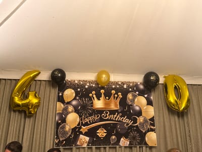 A 40th birthday celebration is evident with a black and gold banner featuring a crown, balloons, and gift illustrations. Gold number balloons "4" and "0" flank the banner. Additional black and gold balloons are also present, some marked with "40." The scene is indoors, with a curtain visible behind the decorations. Part of the ceiling is also visible.