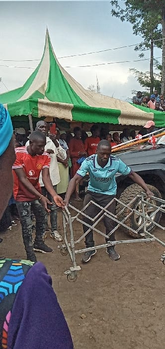 The image shows a group of people standing in front of a large tent and a black vehicle. The coordinates 0.562, 34.805 place the scene in Kivaywa, Kenya, which is located in the Bungoma County.  A man in a blue shirt and black pants is pushing a foldable metal cart towards the vehicle. Another man in a red shirt and black pants is standing on the left side of the cart. They are both wearing black shoes. In the background, there are other people, some of whom are standing and some of whom are seated. The vehicle's tire is visible in the background. The ground is dirt. The sky is overcast.  The image appears to have been taken during a gathering or event, likely in a rural or semi-rural area.  The presence of the foldable metal cart and the vehicle suggests that this could be a gathering for a social or ceremonial event.