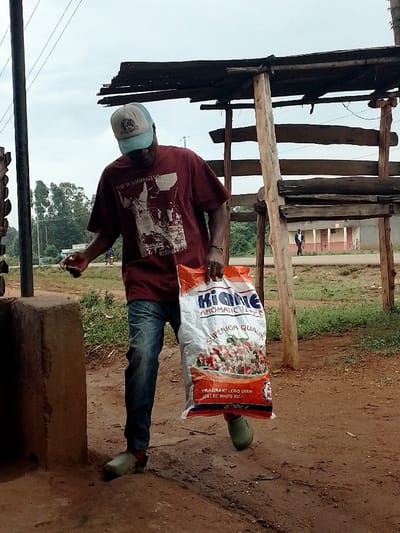 A man wearing a red t-shirt with "The Washington Redskins" printed on it, a blue baseball cap, and blue jeans walks on a dirt path in Kivaywa, Kenya, holding a white bag of rice with "Kionne Aromatic Rice" printed on it. The man is in front of a wooden structure with a thatched roof and an unpaved road. The scene is in a rural area. The man is walking toward the road. The wooden structure appears to be a market stall. There are other people walking in the background. The ground is dusty and brown.  The coordinates provided place the scene within a rural area of Kivaywa.