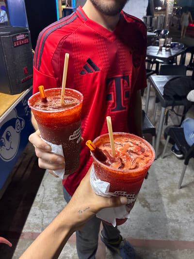 The image depicts an outdoor, nighttime scene in Coatzacoalcos, Mexico, illuminated by artificial light. Two individuals are holding large clear plastic cups containing a dark red, slushy beverage, likely a michelada.
The central figure, partially visible, wears a red Adidas sports jersey with dark blue shoulder stripes and a large "T" logo on the chest. This individual, whose bearded lower face is visible, holds one beverage cup. The cup contains a beige straw and a shorter yellow-and-red chili candy straw, and its rim is coated with a thick layer of red chili powder. Text "LAS MICHELADAS" is partially visible on the cup. A white napkin is wrapped around the base of the cup.
A second person's right arm and hand are prominent in the lower right foreground, holding an identical beverage cup, also with a white napkin, a beige straw, and a yellow-and-red chili candy straw. A small tattoo reading "Te amo" is visible on this person's inner wrist.
In the background to the left, a black speaker box labeled "MAXIMA POTENCIA" and "KAISER" rests on a counter. Adjacent to it is a blue and white sign with a cartoon monkey face. To the right, several dark metal tables and chairs are arranged in an outdoor dining area. Blurry figures, including one person in a light-colored dress, and distant vehicle lights are visible further back, indicating a public street or plaza environment. The ground is a paved surface.