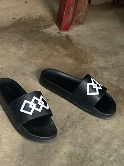 A pair of black slides with a white interlocking geometric design rests on a concrete floor. One slide is positioned in the upper right of the frame, and the other is in the lower right, with the toe of the lower slide pointing towards the bottom left corner. The slides appear to be new or well-maintained. The concrete floor is textured and appears worn in places, with some darker patches suggesting moisture or dirt. In the upper left quadrant of the image, a portion of a red object, possibly a piece of furniture or equipment, is visible, adding a pop of color to the otherwise muted scene. The lighting suggests it is daytime, with no specific indicators of weather conditions. There is no discernible text visible in the image, except for a very faint "GUCCI" logo on the sole of the lower slide, which is mostly obscured. The scene appears to be an indoor or covered outdoor space with a utilitarian or industrial feel, fitting the context of Kuson, Nigeria. No people are present in the image.