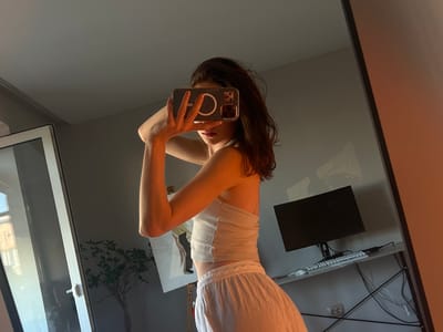 The image is a wide-angle, upside-down, mirror selfie taken indoors. The primary subject is a young woman, who appears to be in her late teens or early twenties, posing for the camera. She is wearing a white, ribbed tank top and white shorts or a skirt. Her dark hair is falling forward, obscuring part of her face. She is holding a smartphone in her right hand, angled to capture the reflection. The phone case is dark with a prominent circular magnetic accessory.
The setting appears to be a home office or study area. In the background, reflected in the mirror, there is a desk with a computer monitor mounted on an arm. A keyboard and various cables are visible on the desk. The walls are a neutral gray, and a large window is visible on the right side of the frame, suggesting it might be daytime. A hanging plant with green leaves is also reflected in the window, adding a touch of nature to the indoor scene. The lighting suggests it might be late afternoon or early evening, with a warm, soft glow, possibly from the sun. The overall mood is casual and relaxed, possibly taken during a moment of leisure at home in Ternopil, Ukraine. There is no visible text in the image.