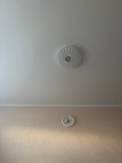 The image shows an interior scene. In the upper portion of the frame, a white, circular light fixture is mounted on a white ceiling. The fixture has a decorative pattern and a central red light. Below the ceiling line, which is demarcated by a beige wall, a white, circular clock is visible on the wall. The clock has Roman numerals and colored hands. The image is blurry and lacks detail, making it impossible to determine specific locations or identify individuals.