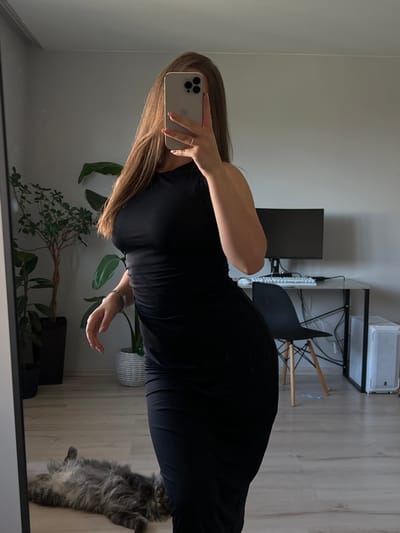 The image shows a woman in a black dress taking a selfie in a mirror. She is holding a silver iPhone in her left hand, with her right hand resting on her hip. Her long, blonde hair is falling over her shoulder. In the background, there is a desk with a computer monitor, keyboard, and mouse. There is also a chair and a cat lying on the floor. The room appears to be a home office or living space. The lighting suggests it is daytime. There is no visible text in the image.