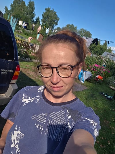 A woman with auburn hair pulled into a ponytail and wearing glasses takes a selfie outdoors. She is wearing a blue t-shirt with a graphic print. Behind her, laundry is hanging on a clothesline. A dark blue SUV is parked to her left. The background features a grassy yard, a wooden fence, trees, and a child's swing set. The overall scene suggests a residential backyard in a rural setting.