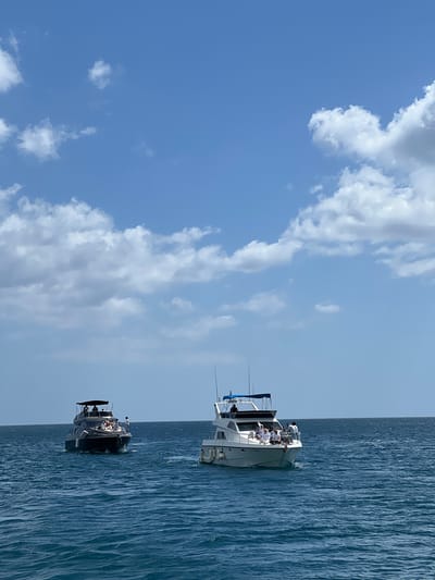 Two boats are sailing on the water under a partly cloudy sky. The location is M'Diq ⵎⴹⵢⴰⵇ المضيق, Morocco. The top boat is white and carries several people. The lower boat is darker in color and also has passengers. The water is blue and rippled.