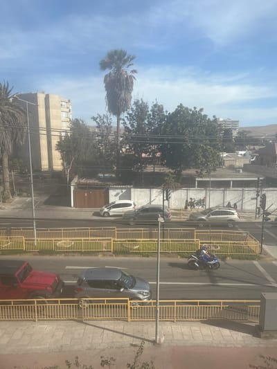 The image depicts a street scene in Arica, Chile, under a bright, cloudy sky. In the foreground, a yellow metal fence borders a sidewalk and a street. On the street, a red Jeep Wrangler is parked to the left, and a silver hatchback is to its right. Further down the street, a person is riding a blue and white motorcycle. Across the street, another yellow fence encloses a grassy area. Beyond the fence, several cars are visible, including a white hatchback and a silver SUV. In the background, there is a multi-story building with a light-colored facade and several trees. A tall palm tree with dry fronds stands prominently near the building. The environment appears to be a residential or mixed-use area in Arica, with buildings and infrastructure indicative of an urban setting.