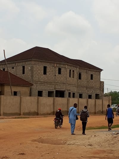 The image depicts a street scene in Keffi, Nigeria, with a partially constructed building as the focal point. The building, made of cinder blocks, has a dark-tiled roof and appears to be two stories high with visible window and door openings. A concrete wall with a rough texture surrounds the property.
In the foreground, the ground is a reddish-brown dirt road. Several people are present:
* A person on a motorcycle, wearing a red cap and dark clothing, is seen riding from left to right.
* Two men are walking from right to left. One is wearing a light blue traditional outfit and carrying a dark bag over his left shoulder. The other is wearing a patterned shirt and black trousers, with a backpack on.
* Further to the right, a young man in a blue tank top and black shorts is walking away from the camera.
* In the background, a white vehicle is parked, and another motorcycle is visible on the right side of the frame.
The sky is overcast with diffused light. The overall environment suggests a developing urban area, possibly a residential or commercial construction site.