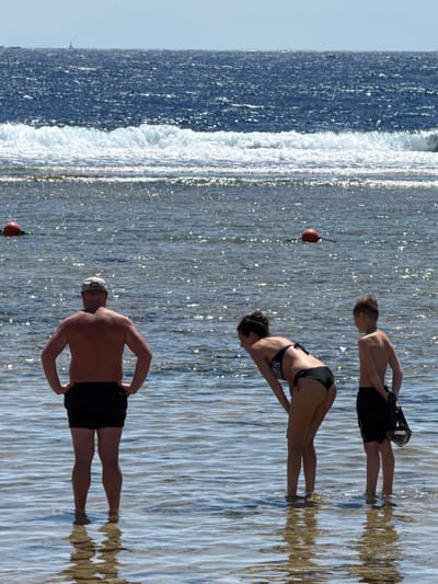 The image depicts three individuals near the shoreline of a body of water, possibly the Red Sea in Sharm El Sheikh, Egypt. The scene is bathed in bright sunlight, suggesting it is daytime, likely mid-morning or afternoon, with clear weather.
In the foreground, a shirtless man wearing black shorts and a cap stands with his hands on his hips, facing away from the camera. His reflection is visible in the rippling water. Behind him, a woman in a black bikini is captured in a dynamic pose, appearing to be in mid-motion, possibly performing an exercise or dive. She is facing away from the camera as well. Further back and to the left, a younger person, likely a boy, is also facing away, wearing black shorts and holding a dark object.
The water is a prominent element, with a line of white surf breaking close to the shore, indicating a shallow area transitioning to deeper water. The surface of the water is highly reflective, creating a shimmering effect due to the sunlight. A red buoy with a black rope is visible floating in the water between the two individuals in the background. The overall atmosphere appears relaxed and leisurely, typical of a beach or coastal resort setting. No visible text is present in the image.