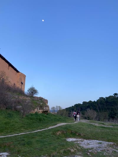 The image shows a path leading away from the viewer, towards a group of three people walking away from the camera, in the direction of a wooded area. The path is dirt and passes through green, grassy terrain, with a rocky outcropping on the left. A tall, tan building, likely part of a historic structure, is visible on the left edge of the image. The sky is a clear, blue, with a single, white moon visible in the upper-middle portion. This scene appears to be on a hillside overlooking the city of Segovia, Spain, located at latitude 40.95, longitude -4.13.