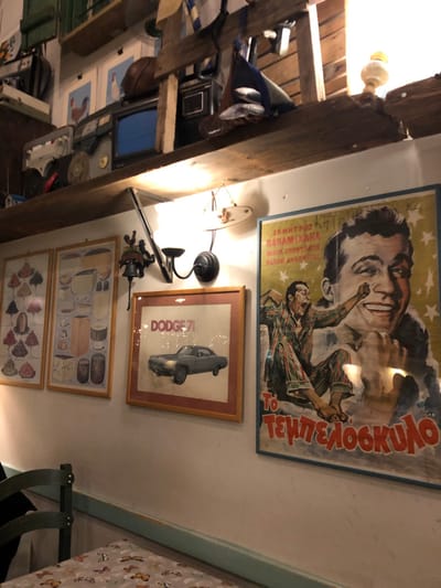 The image captures a warm, eclectically decorated indoor space, likely a room within a home or a establishment in Marousi, Greece. The scene is vertically oriented, featuring a wall adorned with several framed artworks and posters, illuminated by artificial lighting.
Dominating the upper section of the wall is a vintage Greek movie poster for "Ο ΤΕΜΠΕΛΟΚΥΛΟΣ" (The Lazy One), a film starring Dimitris Papamichael. The poster features a large, smiling headshot of Papamichael at the top, and below him, a man in striped pajamas in an animated, jumping pose against a yellow, star-flecked background. The text "ΔΗΜΗΤΡΗΣ ΠΑΠΑΜΙΧΑΗΛ" is also visible.
Below this, a framed poster features a dark-colored car, explicitly identified by the vertical text "DODGE '71" on its left side. Light trails or effects surround the vehicle, suggesting motion. Further down, two more framed illustrations depict collections of food items: the top one showing cheeses, meats, and various kitchen goods, while the bottom one displays an arrangement of smaller items, possibly desserts or pastries.
The room's lighting is warm, with a black wall sconce with a frosted glass shade providing illumination below the movie poster. Another bare light bulb is visible on a large, dark wooden beam that spans the upper left portion of the image, indicating structural elements of the building. A dark, decorative ceiling fan or light fixture hangs below the sconce.
On the left side, behind the wooden beam, a cluttered area includes a blue fabric item, small metallic objects, and a dark electronic device with a screen, possibly an old television. More framed pictures, appearing to be illustrations of birds, are visible below these items. To the right, a portion of a table with a light-colored, patterned tablecloth and a dark chair are visible, suggesting a seating or dining area. The overall atmosphere is cozy and nostalgic, consistent with the cultural cues of the Greek movie poster.
