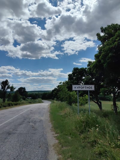 The image depicts a rural road in Ukraine under a bright, cloudy sky. The road is paved but shows significant wear and tear, with patches of cracked asphalt and potholes. To the right of the road, a white rectangular sign with black Cyrillic text is mounted on two metal posts. The text on the sign reads "КУРОРТНОЕ," indicating a place name.
The landscape surrounding the road is lush and green, with abundant trees and tall grass growing along the roadside. A large, mature tree with dark green foliage stands prominently to the right of the sign, casting a shadow. In the background, rolling hills covered in trees and vegetation stretch across the horizon, suggesting a rural or semi-rural setting. The sky is a vibrant blue, interspersed with numerous fluffy white clouds, indicating fair weather. The scene appears to be set during the daytime, likely late morning or early afternoon, given the brightness and the quality of the light. There are no people visible in the image, and the focus is on the natural scenery and the road sign. The overall impression is one of a peaceful, somewhat isolated rural location on a pleasant day.