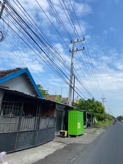 The scene is a street in Kecamatan Tanggulangin, Indonesia (latitude: -7.495, longitude: 112.677). A tall utility pole with multiple electrical wires is on the left side of the road. A building with a blue and gray roof and a metal gate sits to the left of the pole. A green utility box is directly in front of the pole. There is a small tree and some vegetation between the utility box and the road. A motorcycle is traveling on the paved road to the right, with other vehicles visible in the distance. The sky is mostly clear with a few white clouds.