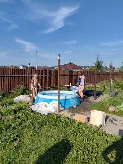 A young boy and a woman are in a backyard with an inflatable pool. The boy, wearing swimming trunks, stands near the pool, looking towards the viewer with a slight smile. The woman, wearing a blue swimsuit and a light pink cover-up, stands next to a lounge chair, looking down at the pool. The inflatable pool is blue and partially filled with water. In the background, a wooden fence encloses the yard, with houses and trees visible beyond. The scene appears to be set on a sunny day, with a blue sky and scattered white clouds, suggesting it is daytime. There are several objects in the yard, including a lounge chair, a bucket, bricks, and some white tarps. The overall mood seems relaxed and summery. The location is identified as Fanipal, Belarus. There is no visible text in the image.