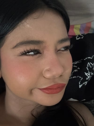 A close-up, eye-level shot focuses on the right side of a young woman's face. Her head is tilted, and her gaze is directed upwards and to her right. She has dark hair that falls to her forehead and right shoulder. Her skin tone is medium, and she is wearing makeup. Her eyebrows are dark and well-defined. Her right eye is open, with long, dark eyelashes. The iris appears to be dark brown. Her left eye is mostly obscured by her head tilt, but a sliver of her iris is visible, also appearing dark. She has prominent cheekbones, with a dusting of blush on her right cheek. Her nose is short and slightly upturned. Her lips are full and painted with a matte, coral-red lipstick. Her chin is slightly turned, creating a subtle dimple. The background is out of focus, with a striped pattern of pink, yellow, purple, and white visible on the left, and a black garment with a white embroidered pattern on the right. The lighting suggests an indoor setting. No specific location in North Jakarta, Indonesia can be determined from the image.