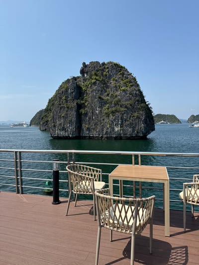 Captured on what appears to be the deck of a ship in Ha Long Ward, Vietnam, this image presents a composition of natural and artificial elements. A prominent limestone karst structure, common to the region, dominates the left side of the frame. It is situated in the body of water, presumably part of Ha Long Bay, that fills the majority of the background. The water extends towards the horizon, visible under a cloudless sky.
To the right, the deck of the vessel takes up a considerable portion of the image. Outdoor furniture is meticulously arranged: a table and chairs made of a light-colored material are staged, seemingly for relaxation or dining. A railing separates the seating area from the water. Below the railing, there's a buoy.
The lighting suggests a bright, sunny day, casting sharp shadows on the deck's surface and illuminating the textures of the rock formation. The composition is vertical, emphasizing the height of the karst and the depth of the scene.
