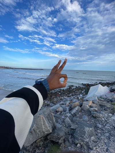 The scene is an outdoor shot, likely taken from a beach, featuring a person's hand making a gesture in the foreground. In the background is the beach and sea and along the coast is trash. The sky above is filled with white clouds against a blue sky. The location is Alaguntan, Nigeria.