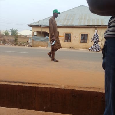 The image depicts a street scene in Tempure, Nigeria. The foreground shows a dusty reddish-brown surface, likely a road or pathway, with a concrete structure bordering it. In the midground, a man wearing a brown traditional outfit and a green baseball cap walks along the street, carrying a cylindrical object. He is wearing sandals. Behind him, a woman in a patterned dress and headscarf is also walking, appearing to move away from the viewer.
The background features a building with a corrugated metal roof and light-colored walls. There is some graffiti or writing on the wall of the building, including "AZOA SAYA RUWA I" and "+425" near a window, and "10.7M" with a circle symbol further to the right. In the distance, other buildings and vegetation are visible under an overcast sky, suggesting it might be a cloudy day. The overall impression is of an everyday street scene in a Nigerian town. The activity taking place is people walking along the street. There are no clear indicators of a specific event.