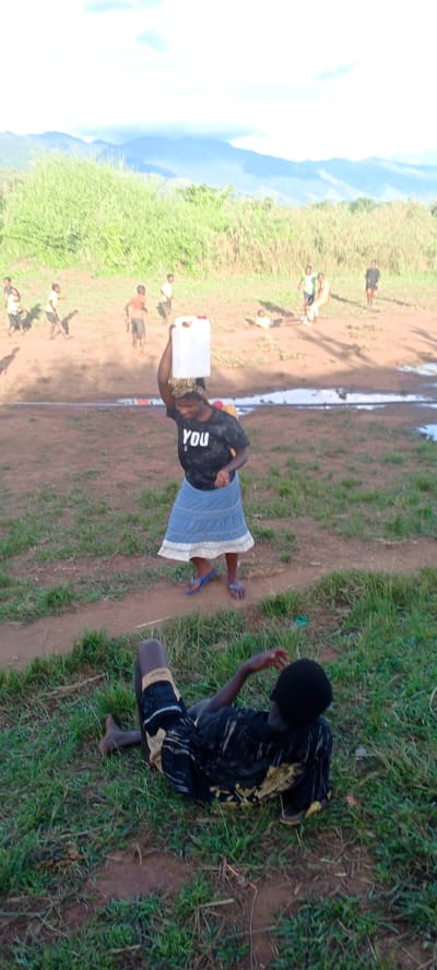 In a daytime shot, a woman in Ngalula, Democratic Republic of the Congo, is balancing a white plastic container on her head, wearing a black t-shirt with the word "YOU" printed on it, and a blue skirt with a white trim. She is wearing blue sandals. In the foreground, a person is lying on the grass. In the background, several people are on a field with a muddy area. The background also features a grassy hill and distant mountains under a partly cloudy sky.