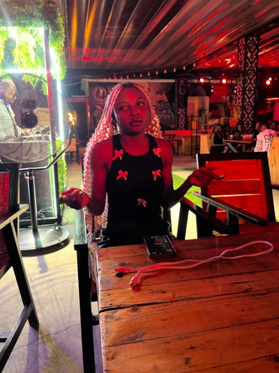 The media file shows a young woman sitting at a wooden table outdoors, possibly at a restaurant or bar. She has braids and is wearing a black top with red bows. Her left hand is raised, and she appears to be looking towards the camera. A smartphone and a red charging cable are on the table next to her.
In the background, there are red lights, decorative metalwork, and plants. A large fan is visible in the lower left corner of the image. The scene is illuminated by artificial lights, suggesting it might be evening or nighttime. The overall atmosphere appears casual and relaxed. There is no visible text in the image. The location context is Howayi, Nigeria.