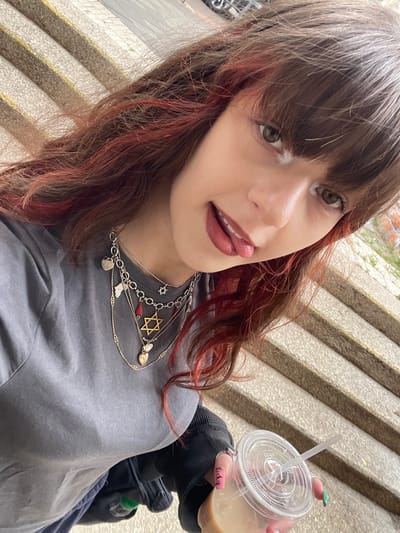An upward-angled, close-up photograph depicts a young adult female in the city of Raanana, Israel, captured in a selfie-style pose. The subject possesses medium brown hair with prominent reddish-pink highlights, styled with straight bangs across her forehead. Her brown eyes are open, and she is looking directly at the camera with a slight smile and her tongue subtly protruding from her mouth, which is adorned with dark reddish-brown lipstick.

She is wearing a plain grey short-sleeved t-shirt. On her neck, she displays multiple layered necklaces featuring various pendants: a large gold-toned Star of David, a smaller silver-toned Star of David, several heart-shaped charms (both silver and gold-toned), a small silver bird or dove, and a red drop-shaped charm. Her right hand is visible, clad in a dark long-sleeved garment, holding a clear plastic cup with a lid and a white straw. The cup contains a light brown liquid, consistent with an iced coffee or similar beverage. Her fingernails are painted, with visible colors including pink/red and green, and part of the word "Lucky" is inscribed on one nail.

The background consists of light-colored concrete or stone steps with horizontal indentations, suggesting an outdoor setting like a staircase or tiered seating. Faint hints of greenery are visible to the far right, and indistinct urban elements can be perceived in the upper background. The lighting suggests natural daylight.