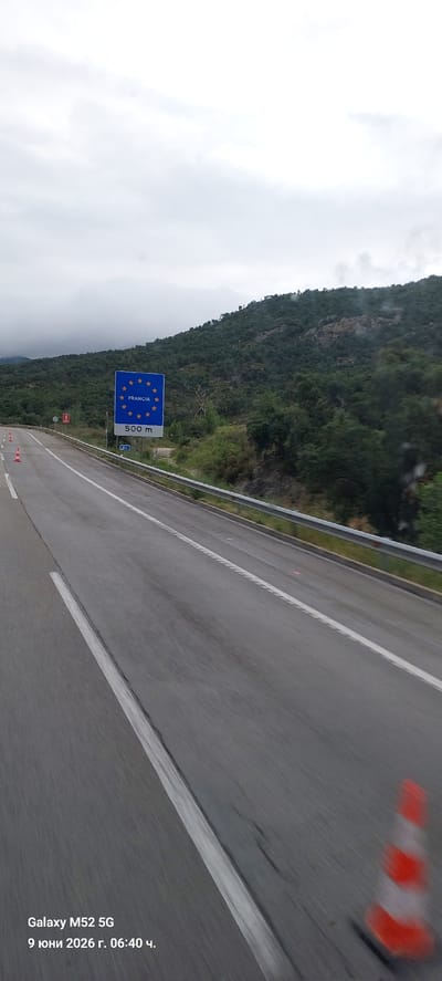 The image captures a view from a vehicle traveling on a highway, likely near the border. In the foreground, the asphalt road is visible with white lane markings. On the right side of the road, a section of a metal guardrail runs parallel to the highway. Further to the right, a blurry orange and white traffic cone is present. In the middle ground, a blue sign with a ring of yellow stars, characteristic of the European Union, is mounted on a pole. Below the stars, the word "FRANCIA" is written in white letters, indicating France. Beneath "FRANCIA", "500 m" is displayed, suggesting the distance to the French border. To the left of the France sign, a smaller red and white sign is visible on a pole. The background features a densely forested hillside under a cloudy, overcast sky. The text in the bottom left corner reads "Galaxy M52 5G 9 юни 2026 г. 06:40 ч.", indicating the device used to capture the image and the date and time, which is likely a timestamp from a mobile device. This scene is consistent with the location of the border crossing between Spain and France, near the town of La Jonquera, Spain, which is situated on the AP-7 highway.