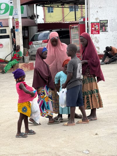 This image, taken in Lagos, Nigeria, depicts a group of people standing in what appears to be a roadside area, possibly near a gas station or market. The subjects include several women and children, all of whom are looking in the same direction, away from the camera.
The people are dressed in vibrant, patterned clothing, with many of the women wearing headscarves. One woman in the foreground is wearing a bright pink top and a yellow skirt with a blue flower pattern, accessorized with orange beads. Another woman is wearing a light pink headscarf and a traditional patterned dress. A child in the middle is wearing a grey t-shirt and blue shorts, carrying a white bag.
The background shows a building with advertisements on its wall, including one for Coca-Cola. There are also what look like refrigerators or freezers inside the building. A silver car is parked behind the people. A gas pump is visible in the lower part of the image, with a man in a green and red shirt standing next to it. The ground is paved with concrete.
The lighting suggests it is daytime, and the weather appears to be fair. The overall mood of the scene is one of stillness, with the subjects appearing to be waiting or observing something off-camera. There is no explicit indication of a specific event, but the gathering of people and the presence of goods suggests a commercial or social interaction.
Visible text includes "75 NAIRA" and a Coca-Cola logo on an advertisement. There are also signs and logos on the gas pump and in the background that are not clearly legible.