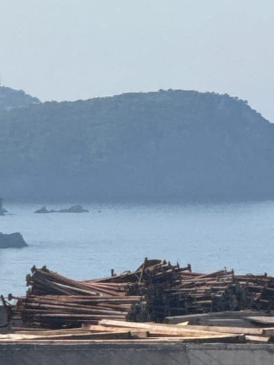 This is a vertical, slightly blurry, close-up photograph taken in Boreti, Montenegro. The image features two main subjects: a vast, pale blue body of water occupying the majority of the frame, and a construction scaffold on the right side of the image, composed of numerous wooden poles. The water appears calm and reflective, with a slight haze that softens the distant horizon.

The left side of the image is dominated by a soft, out-of-focus mountainous landscape, rendered in muted shades of blue and gray. The shape of the mountain suggests a natural, possibly coastal, environment. The lighting is soft and diffused, indicating it might be a hazy day or early morning/late afternoon.

The construction scaffold on the right is detailed, showing an arrangement of vertical and diagonal wooden poles, some of which appear to be bound together. The texture of the wood is visible, and the structure seems to be unfinished or in the process of being erected. The contrast between the natural elements of water and mountain and the man-made structure of the scaffold is a prominent feature of the image.

No people are visible in the frame. The overall impression is one of a serene natural setting juxtaposed with ongoing construction. The time of day is likely during daylight hours, but the exact time is difficult to determine due to the lighting conditions. No text is visible in the image.