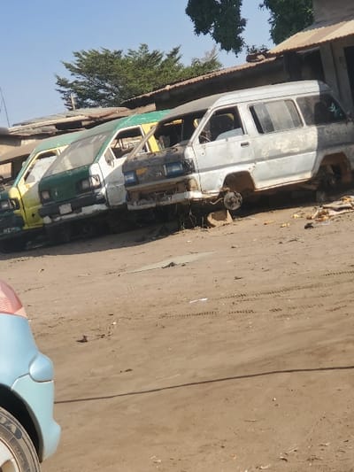 This image displays a vehicle salvage or long-term storage yard situated in Kaduna South, Nigeria, under bright daylight. The foreground reveals the partial left front fender and tire of a light blue vehicle. Dominating the midground are three abandoned passenger vans, parked in a linear arrangement on an unpaved, reddish-brown dirt lot exhibiting tire tracks and minor debris. From left to right, a yellow van and a green van are visible, both appearing dirty and worn. To their right, a heavily stripped gray/silver Toyota passenger van is present; its front bumper, headlights, and all four wheels are missing, and it rests on visible support blocks. The interior of this gray van is exposed and appears disassembled. In the background, low-rise structures with corrugated metal roofs (brown and green) are discernible, topped by sparse green trees against a clear blue sky. No individuals or specific actions are observed within the scene.
