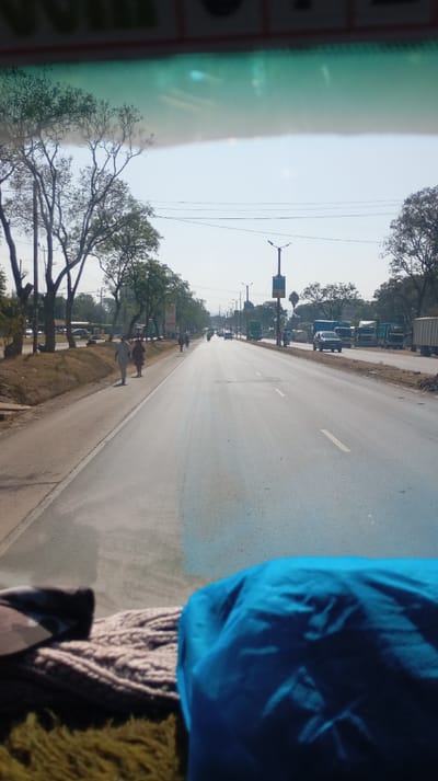 A view from inside a vehicle, looking out onto a wide, multi-lane road. Trees line the side of the road, and pedestrians walk on the shoulder. Various vehicles, including cars and trucks, travel in both directions. Light poles and signage are visible along the roadside. A blue item sits in the foreground within the vehicle's cabin. The sky is bright, suggesting daytime.