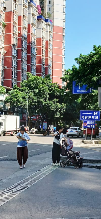 The image depicts a street scene in the Jiulongpo District of Chongqing, China. A tall, multi-story building with red and white facades and numerous windows dominates the left side of the frame. Trees with dense green foliage line the street, partially obscuring signs written in Chinese characters.

On the sidewalk, a woman in a white shirt and black pants stands next to a stroller. Further down the sidewalk, another woman holds a baby in her arms. Both women appear to be on the sidewalk, with a metal grate bordering the sidewalk.

Vehicles, including a white car and a white truck, are visible on the street. The sky is clear and blue.