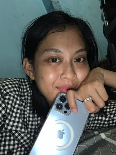 An individual is depicted indoors in Kecamatan Pati, Indonesia, captured in a close-up, eye-level shot. The subject, a female, has dark hair, dark eyes, and a slight smile, looking directly at the camera. She wears a black and white checkered shirt. Her right hand, adorned with a multi-stone ring on the ring finger, rests near her chin and mouth, holding a light blue smartphone. The phone, identifiable as an iPhone with three rear camera lenses, is encased in a clear protective cover displaying a white MagSafe ring centered on the Apple logo. The background consists of a light blue wall section to the upper left and indistinct dark objects or furniture elements to the upper right, consistent with an interior setting, possibly a bedroom.