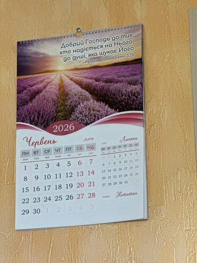 This is a close-up, slightly angled shot of a wall calendar. The calendar features a scenic photograph of a vibrant lavender field at sunrise or sunset, with golden light filtering through the sky. The top of the calendar has text in Ukrainian, which translates to "The Lord is good to those who hope in Him, to the soul that seeks Him. Bible. Lamentations of Jeremiah 3:25". The calendar itself is open to the months of June and July of the year 2026. Each month has a grid for daily entries with days of the week labeled in both Ukrainian and English. The Ukrainian days are: ПН (Monday), ВТ (Tuesday), СР (Wednesday), ЧТ (Thursday), ПТ (Friday), СБ (Saturday), and НД (Sunday). The English translations are also provided below. The calendar is bound by a silver spiral binding. The background is a textured, light brown surface, possibly a wall.