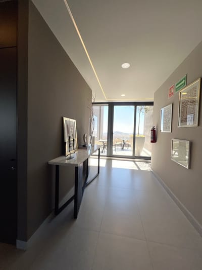The image displays a modern, well-lit interior hallway or corridor situated in León, Mexico. The perspective is directed towards the end of the hallway, where dark-framed double glass doors open onto an outdoor terrace with a panoramic view of rolling hills under a clear blue sky.
The left wall is finished in a dark brown or grey-brown textured material. A dark, possibly solid wood, door is partially visible on the far left. A contemporary console table with a white marble top and a black metal base, featuring vertical slatted details, is positioned against this wall. Adorning the console table are a gold-framed artwork, a black metal candelabra, a tall clear vase containing decorative branches, and other indistinct decorative items.
The right wall is a light beige or greige hue with a subtle pattern. Mounted on this wall are a red square sign with white iconography, a green rectangular "EXIT" sign with a white arrow pointing right, a red fire extinguisher, and a cluster of four framed items. The upper two frames display abstract or geometric artwork in earthy tones, while the lower two are smaller, one appearing to be a mirror or highly reflective framed image.
The floor consists of large, light-colored square tiles, reflecting the natural light streaming in from the glass doors. The white ceiling incorporates a linear LED light strip running along the left wall's edge and several circular recessed lights.
Beyond the glass doors, the outdoor terrace area is furnished with at least two round patio tables and associated chairs, alongside a large white planter containing a small tree. No individuals are present, and no actions or interactions are depicted within the scene.