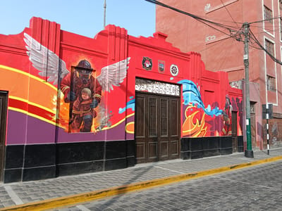 The image depicts a building facade with murals in Barranco, Peru. The building is predominantly red with black accents at the base. There are two large murals: one featuring a firefighter with wings carrying a child, and another showing a firefighter with a hose. The central portion of the facade includes a large wooden door with decorative metalwork and two emblems above it. The sidewalk in front of the building is paved with bricks and a yellow curb. A utility pole with a complex array of wires is located to the right of the building. An adjacent building, painted in a lighter shade of red, is visible behind the facade.