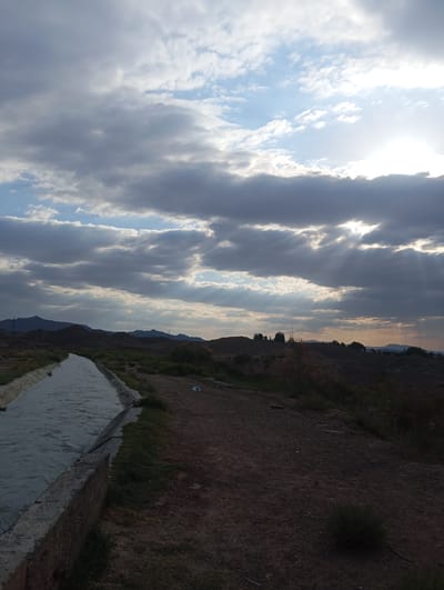 This is a vertical, medium-shot photograph of an irrigation canal in a rural area. The canal, which is concrete-lined, runs diagonally from the lower left corner towards the center of the image. It is filled with dark, rippling water. To the right of the canal is a dirt path, with sparse green vegetation growing along its edge and on the embankment beyond. In the distance, silhouetted against a dramatic sky, are rolling hills and mountains. The sky is filled with a mix of dark, heavy clouds and patches of brighter blue, with shafts of sunlight breaking through, creating a crepuscular effect. There are no people visible in the image. The overall impression is one of a vast, somewhat desolate landscape under a dynamic sky. The location is specified as Kızıl-Bel, Kyrgyzstan.