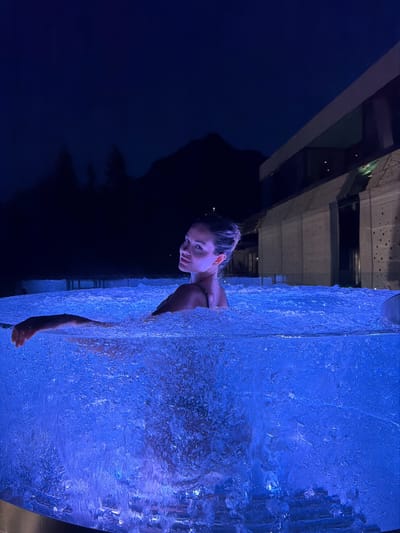 The image shows a woman in a hot tub at night. The hot tub is filled with blue, bubbling water, and the surrounding area is dark. In the background, there are mountains and a modern building. The woman is looking over her shoulder at the camera, with a slight smile on her face. The scene appears to be located in San Pellegrino Terme, Italy.